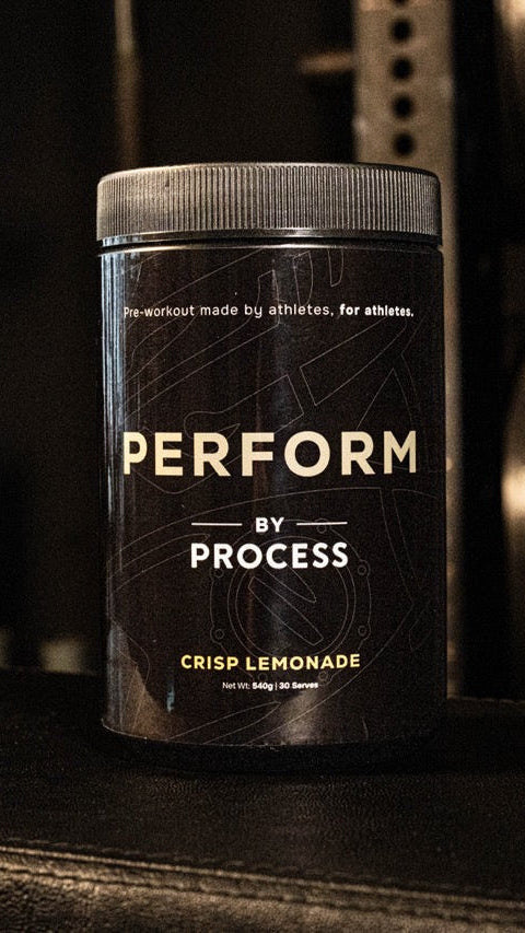 A black tub of Process Performance supplement in Crisp Lemonade flavor, standing on a dark surface in a gym setting with weight racks blurred in the background.