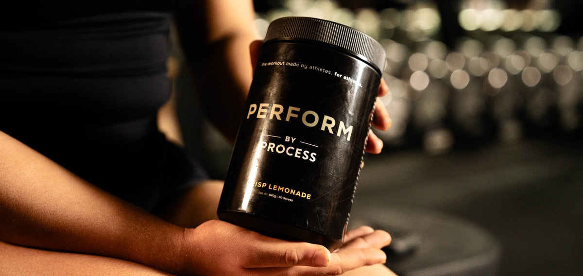 Person holding a 'PERFORM by PROCESS' supplement container in a gym setting